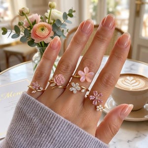Può includere: Una mano mostra cinque anelli d'oro, ognuno ornato da un motivo floreale. Gli anelli presentano pietre preziose rosa e trasparenti, con una varietà di forme di fiori. Una tazza di caffè e un mazzo di fiori sono sullo sfondo.