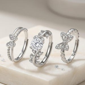 May include: Three silver rings with intricate designs. One ring features a large, round, clear stone, while the others have heart and bow shapes adorned with smaller stones. The rings are displayed on a white surface.