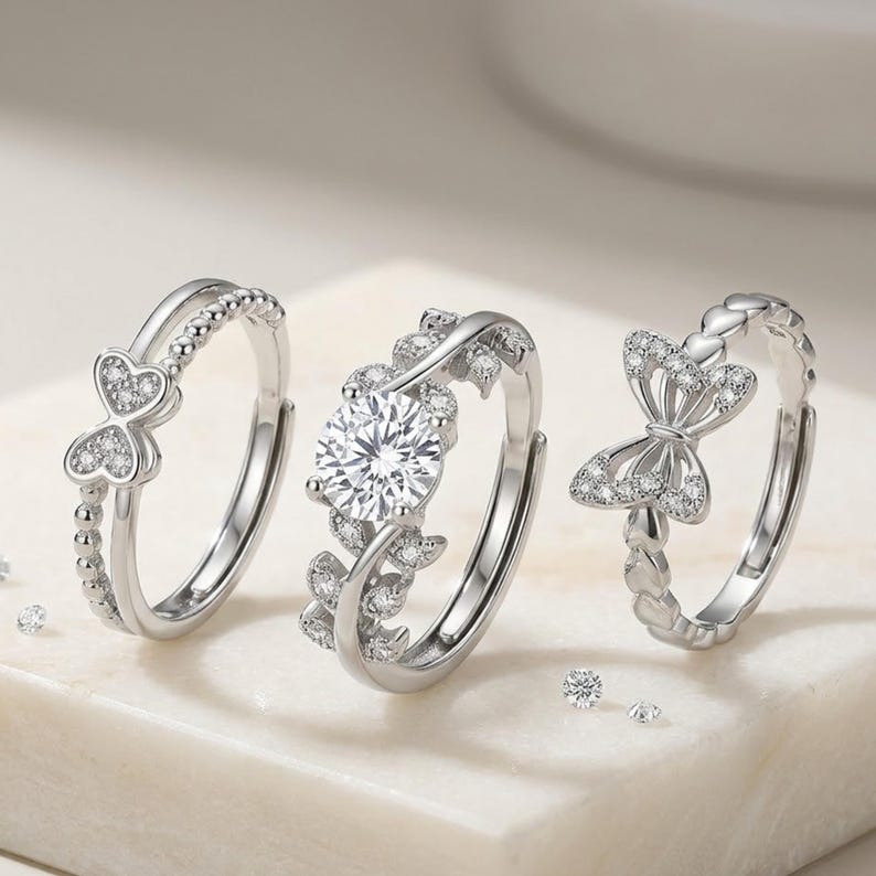 May include: Three silver rings with intricate designs. One ring features a large, round, clear stone, while the others have heart and bow shapes adorned with smaller stones. The rings are displayed on a white surface.