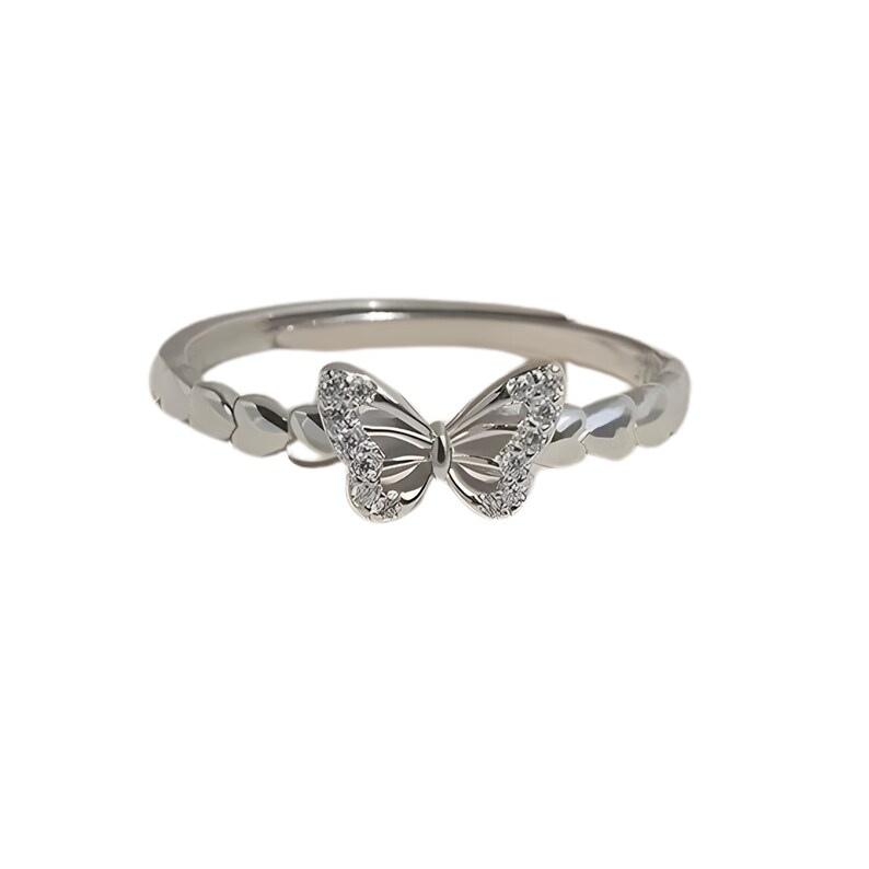May include: A silver-colored ring featuring a butterfly design. The butterfly has a detailed wing pattern and is embellished with small, clear gemstones. The band has a textured design. The ring is set against a white background.