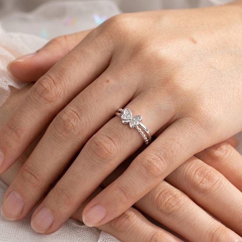 May include: A silver ring featuring a butterfly design with clear gemstones. The ring has a double band with a beaded texture on the lower band. The butterfly is the focal point, with the wings adorned with small, sparkling stones.
