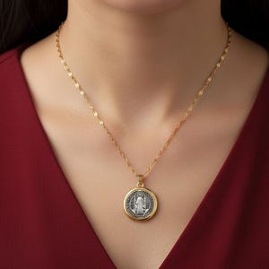 Può includere: Una collana color oro con un ciondolo rotondo. Il ciondolo presenta un'immagine religiosa color argento all'interno di una cornice color oro. La collana è indossata su un capo rosso.