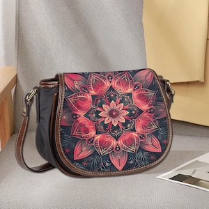 以下が含まれることがあります： 黒いサドルバッグ、茶色の革のストラップとトリム付き。 フラップには、赤、ピンク、青の色合いの鮮やかなマンダラデザインが施されています。 バッグは曲線的な形状で、金色のバックルが付いています。