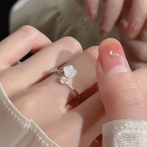 May include: A silver ring with a white rose and a clear gemstone. The ring wraps around the finger, with the rose and gemstone on either side. The ring is displayed on a hand with manicured nails. The ring is a delicate piece of jewellery.