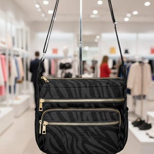 Pode incluir: Bolsa transversal preta com padrão de listras de zebra. A bolsa tem três compartimentos com zíperes dourados e uma alça longa e ajustável. A bolsa é exibida em um ambiente de loja de varejo.