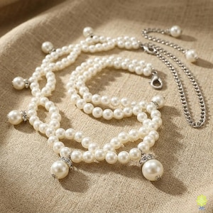 Puede incluir: Un collar de varias hileras con perlas blancas artificiales y detalles en metal plateado. El diseño incluye bucles de perlas interconectados con dijes de perlas colgantes, cada uno engastado en un anillo plateado con cristales claros.