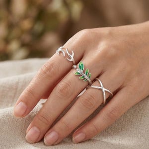 Pode incluir: Uma mão exibe quatro anéis de prata. Um anel apresenta um design de chifre de veado, outro tem uma folha verde e branca, e um terceiro é um design simples em forma de cruz. Os anéis estão em uma mão com um fundo neutro.