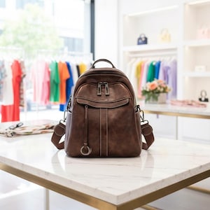 May include: A brown leather backpack with a top handle, front pocket, and adjustable shoulder straps. The backpack has silver-coloured zips and a decorative ring. The bag is on a white marble table.