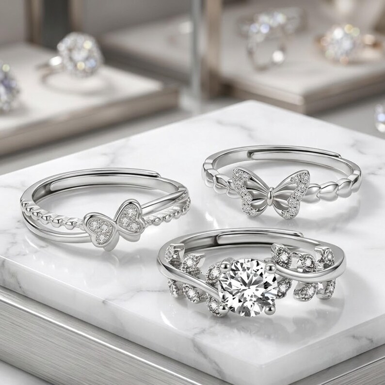 May include: Three silver rings with clear gemstones. One ring features a butterfly design, another has a large round stone, and the third has a double band with a butterfly. The rings are displayed on a white marble surface.