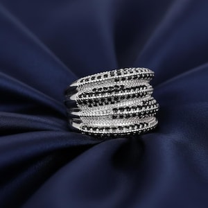 Può includere: Un anello d'argento con più fasce, ognuna ornata da file alternate di pietre nere e trasparenti. L'anello è posto su uno sfondo blu scuro e setoso, che ne evidenzia il design intricato e la brillantezza.