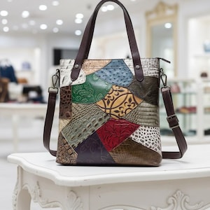 Puede incluir: Un bolso tote de cuero patchwork multicolor con un asa de cuero marrón y una correa de hombro desmontable. El bolso presenta varias texturas y colores, incluyendo verde, marrón, rojo y blanco. El bolso está sobre una mesa blanca.