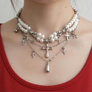 Puede incluir: Un collar de varias capas con perlas de imitación y abalorios plateados. El collar tiene tres hilos de perlas blancas de imitación, con cruces plateadas, estrellas y un colgante en forma de lágrima. El collar se lleva sobre una camiseta roja.