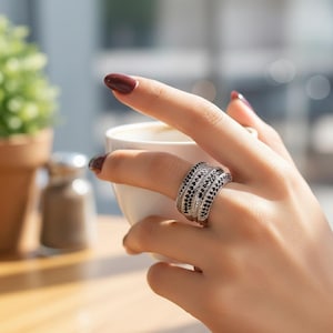 Può includere: Un anello argentato a più fasce, ognuna ornata da pietre nere e trasparenti alternate. L'anello è indossato su un dito. La mano tiene una tazza bianca, con uno sfondo sfocato.
