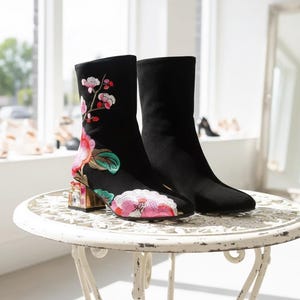 Puede incluir: Botines negros con bordado floral. Las botas presentan un diseño colorido de flores y hojas rosas, rojas y verdes. Los tacones son de bloque transparente con detalles dorados. Las botas están sobre una mesa blanca ornamentada.