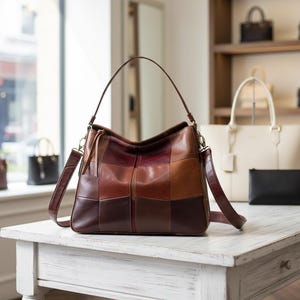 Peut inclure: Sac à bandoulière en cuir marron et bordeaux, avec un motif patchwork. Il est posé sur une table blanche. On aperçoit d'autres sacs en arrière-plan, sur des étagères et un rebord de fenêtre.