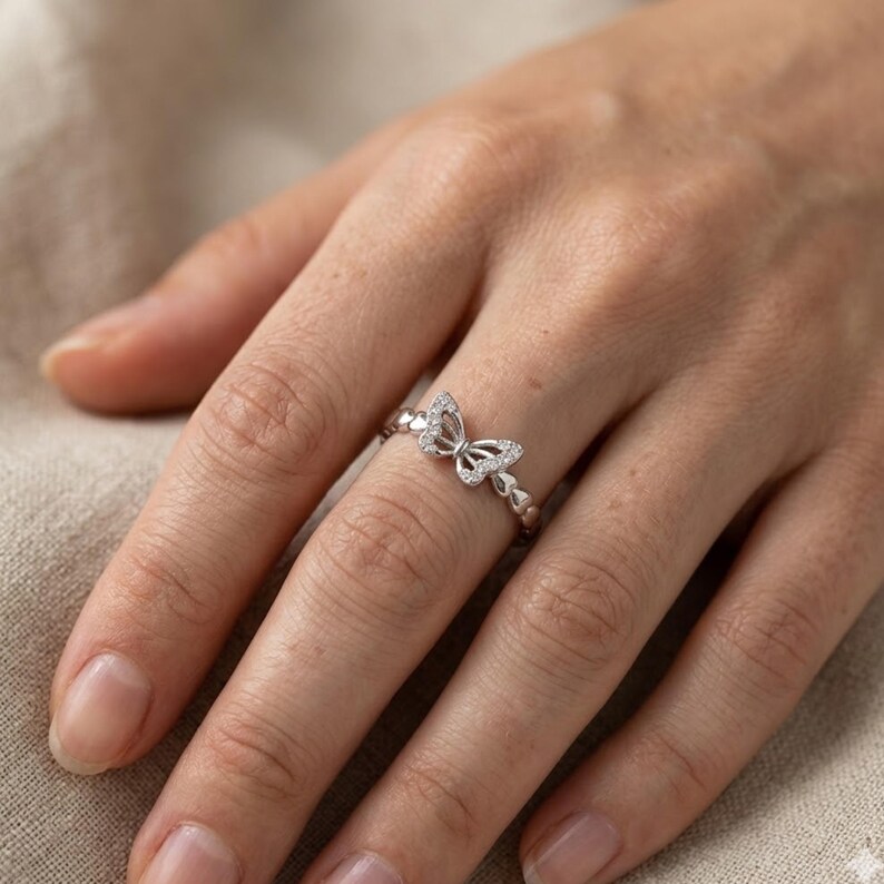 May include: A silver butterfly ring with small, clear gemstones. The butterfly is the focal point, with delicate wings and a detailed body. The band features small heart-shaped accents. The ring is worn on a finger.