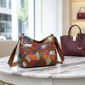 Può includere: Una borsa a tracolla patchwork multicolore con cinturini e cerniere marroni. La borsa presenta un motivo geometrico nei toni del blu, rosso, arancione e bianco. La borsa è appoggiata su un tavolo di marmo.