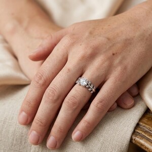 May include: A silver ring with a large, round, clear gemstone at the center. The band features a leaf design with smaller, clear gemstones. The ring is on a hand with a neutral background.