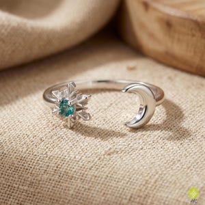 Puede incluir: Anillo plateado con diseño de copo de nieve con gema turquesa y media luna. El anillo se muestra sobre un fondo neutro, resaltando los detalles intrincados de la joyería.