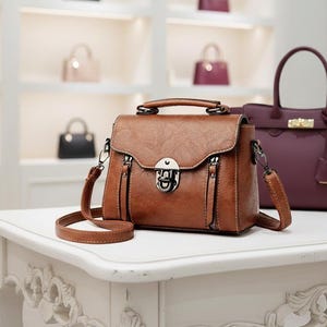 Pode incluir: Bolsa de couro marrom com uma alça superior e uma longa alça ajustável. A bolsa tem uma superfície texturizada, um fecho prateado e dois zíperes frontais. Outras bolsas estão visíveis no fundo.