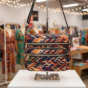 Może przedstawiać: Torba crossbody z kolorowym wzorem fal w odcieniach pomarańczu, zieleni, niebieskiego i fioletu. Torba posiada wiele przegródek zapinanych na zamek błyskawiczny ze złotymi zamkami i czarny pasek na ramię. Torba jest prezentowana na białej powierzchni.