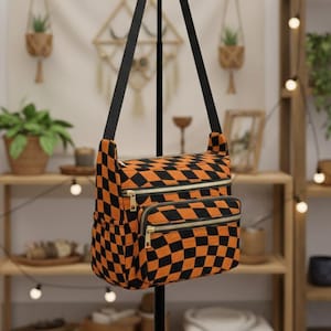 May include: An orange and black checkered crossbody bag with a black strap and gold zippers. The bag has two zippered compartments and a side pocket. The bag is made of a soft, textured fabric.