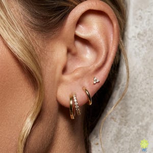 May include: Close-up of an ear adorned with gold earrings. The ear has three piercings, each with a different style of earring. One piercing features a small, gold stud with three small stones. The second piercing has a small gold hoop. The third piercing has a gold hoop with small stones.