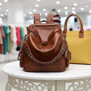 Può includere: Uno zaino in pelle marrone con borchie argentate sulle spalline. Lo zaino ha più tasche e una maniglia superiore. Una borsa tote gialla è visibile sullo sfondo.