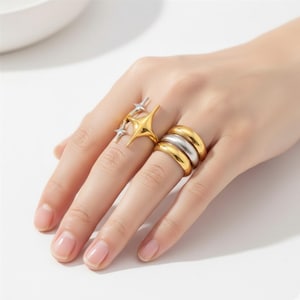 Puede incluir: Una mano muestra tres anillos. Uno presenta un diseño dorado de estrella con detalles plateados. Otro anillo tiene tres bandas apiladas, dos doradas y una plateada. Los anillos están sobre un fondo claro.