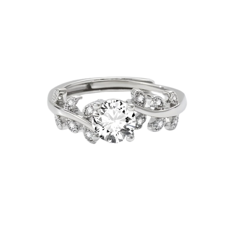 May include: A silver-toned ring featuring a large, round, clear gemstone at the center. The ring is adorned with smaller, clear gemstones and leaf-like designs on the band, creating an elegant and nature-inspired aesthetic.