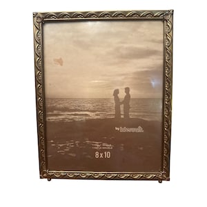 May include: A rectangular gold-colored picture frame with an ornate, scalloped edge. The frame holds an 8 x 10 inch photograph of a couple on a beach. The frame has a small stand and the text "by Intercraft".