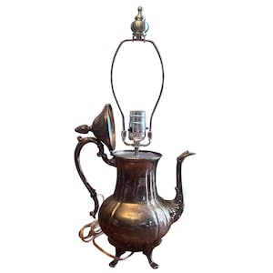 May include: A silver-toned metal teapot lamp featuring a curved handle, a spout, and a hinged lid that is propped open. The lamp includes a central light socket and a metal harp assembly extending upward, with a power cord visible at the base.