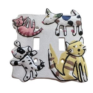 May include: A white double light switch plate featuring a textured surface and hand-painted illustrations of four animals. The design includes a pink cat, a spotted dog, a black and white dog, and a yellow cat, all outlined in thick black lines.