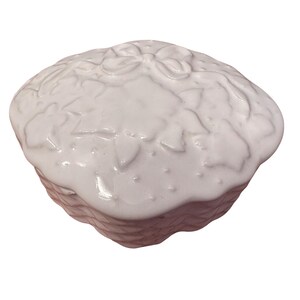 May include: A white ceramic trinket box with a lid. The lid features a raised floral and bow design. The box has a basket weave pattern on the sides. The box is round and has a glossy finish.