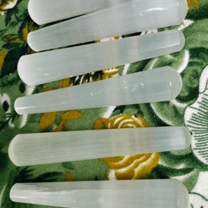 May include: Six cylindrical selenite wands with a translucent, white, fibrous appearance and rounded ends. The wands are arranged in a parallel formation, showcasing their smooth, elongated shapes and natural mineral texture in a clear, bright light.
