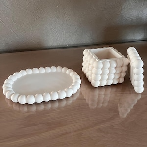 Op de afbeelding: Een set van drie crèmekleurige keramische items: een ovaal dienblad met een kralenrand, een vierkante doos en een deksel. De doos en het deksel hebben een textuur met een bubbelachtig ontwerp. De items zijn gerangschikt op een houten oppervlak.