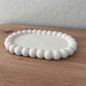Puede incluir: Una bandeja decorativa blanca de forma ovalada con un borde de cuentas. La bandeja está hecha de un material sólido y opaco y se encuentra sobre una superficie de madera. El borde de cuentas está compuesto por pequeñas cuentas redondas y espaciadas.