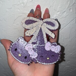 Puede incluir: Un charm en forma de Hello Kitty morado con un lazo grande y dos lazos más pequeños. El charm tiene una superficie texturizada y detalles blancos. El charm se sostiene en una mano.