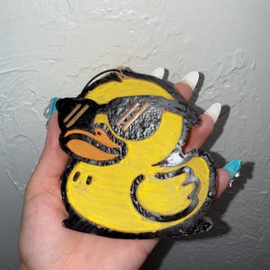 May include: A yellow rubber duck ornament featuring black outlines and black sunglasses. The duck has an orange beak and a textured surface finish. A thin string is attached to the top of the duck for hanging purposes, and the design is flat.