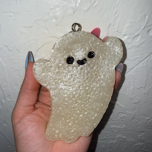Puede incluir: Adorno en forma de fantasma de color claro, con una cara sonriente y un lazo metálico para colgar. El fantasma tiene una superficie texturizada y se sostiene en una mano sobre un fondo blanco. Para Halloween.