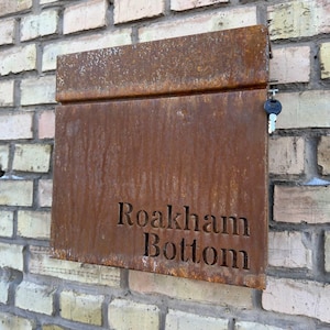 Op de afbeelding: Een rechthoekige, verweerde metalen brievenbus met de woorden "Roakham Bottom" in de voorkant uitgesneden. De brievenbus heeft een roestbruine kleur en is bevestigd aan een bakstenen muur. Een sleutel hangt aan de zijkant.