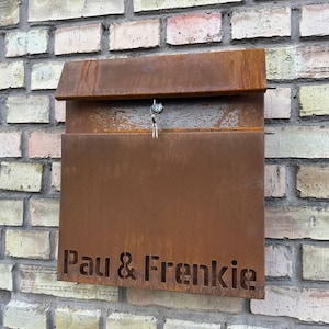 May include: A weathered, rust-colored metal mailbox mounted on a brick wall. The mailbox has a hinged top and the names "Pau & Frenkie" are cut out on the front. A key hangs from the lock.