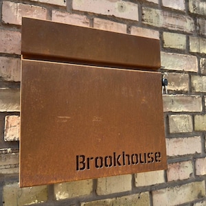 May include: A rectangular, rust-coloured metal post box featuring a horizontal mail slot at the top and the word Brookhouse cut out in a stencil-style font at the bottom right. A small silver key is inserted into a lock on the right side edge.