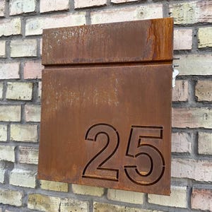 Può includere: Una cassetta postale rettangolare in metallo usurato con il numero 25 intagliato. La cassetta postale è di colore marrone arrugginito ed è montata su un muro di mattoni. Il muro di mattoni ha una miscela di mattoni marroni chiari e scuri.