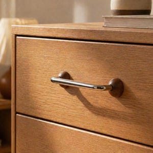Puede incluir: Primer plano de una cómoda de madera con tres cajones. Cada cajón tiene un tirador plateado con un pomo de madera marrón oscuro. La veta de la madera es visible y la cómoda tiene un tono cálido y natural.