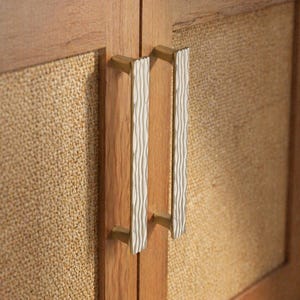 May include: Close-up of a wooden cabinet with woven panels and gold-toned hardware. The cabinet doors feature vertical, textured white and gold handles. The cabinet's frame is a light brown color.