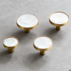 Può includere: Quattro pomelli per mobili con una faccia rotonda in madreperla bianca e una base dorata. I pomelli sono disposti su una superficie grigia. I pomelli variano leggermente di dimensioni, il più grande misura circa 5 cm di diametro.