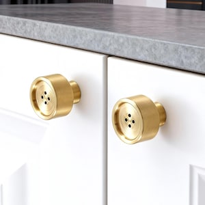 May include: Two brass cabinet knobs with a button-like design. Each knob features a circular face with four small holes and a cylindrical base. The knobs are mounted on a white cabinet door.
