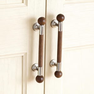 May include: Two cabinet door pulls with a dark brown wooden handle and a silver metal accent. The pulls have a round wooden ball at each end. The pulls are mounted on a cream-colored cabinet door.