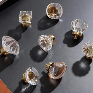 Puede incluir: Una colección de pomos de armario transparentes y dorados en varias formas, incluyendo diseños redondos, cuadrados y facetados. Los pomos tienen una base de latón y están dispuestos sobre una superficie oscura.